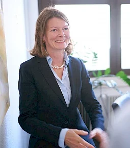 Eine Frau in einem dunklen Anzug und Perlenkette, die lächelt und sich mit einer anderen Person unterhält.