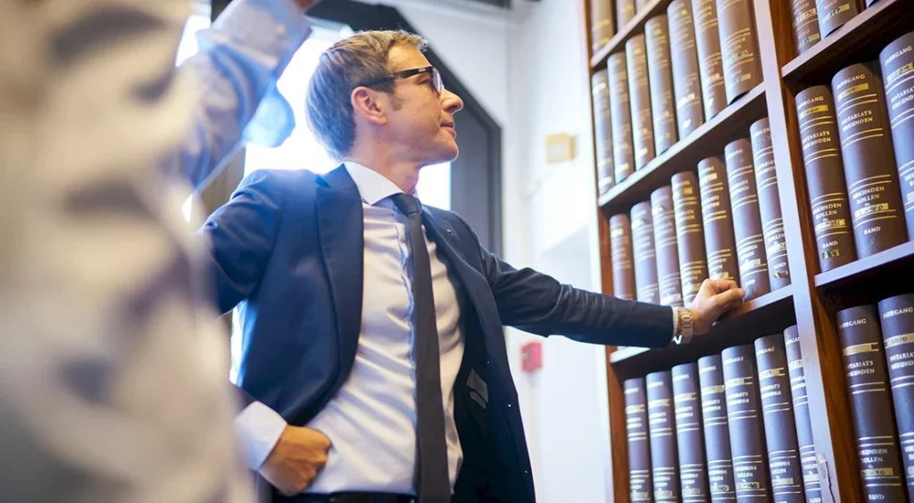 Ein Anwalt steht in einer Bibliothek neben Regalen mit Gesetzesbüchern und betrachtet sie.