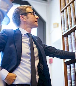 Mann in Anzug mit Brille steht vor einem Bücherregal und zeigt auf die Bücher.