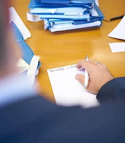Eine Person, die mit einem Stift auf ein Blatt Papier schreibt, während Aktenstapel im Hintergrund sichtbar sind.