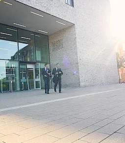 Zwei Männer in Anzügen gehen vor einem modernen Gebäude mit großen Fenstern.